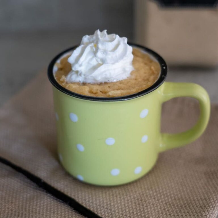 A yellow polka dot mug filled with a mug cake, topped with a swirl of whipped cream, sits on a textured surface.