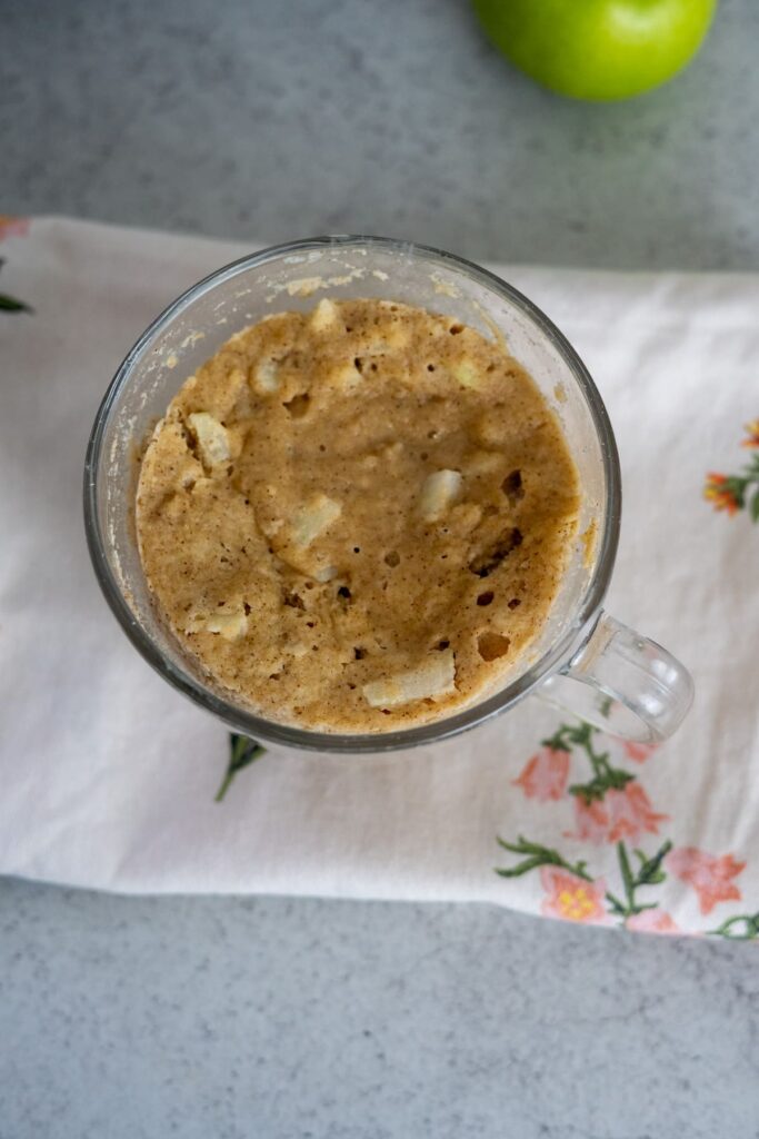 A glass mug filled with a keto apple cinnamon mug cake containing small apple pieces, placed on a white floral napkin with a green apple in the background.