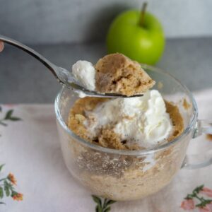 A clear glass mug with a baked dessert topped with whipped cream; a fork holds a bite above the mug. A green apple is in the background.