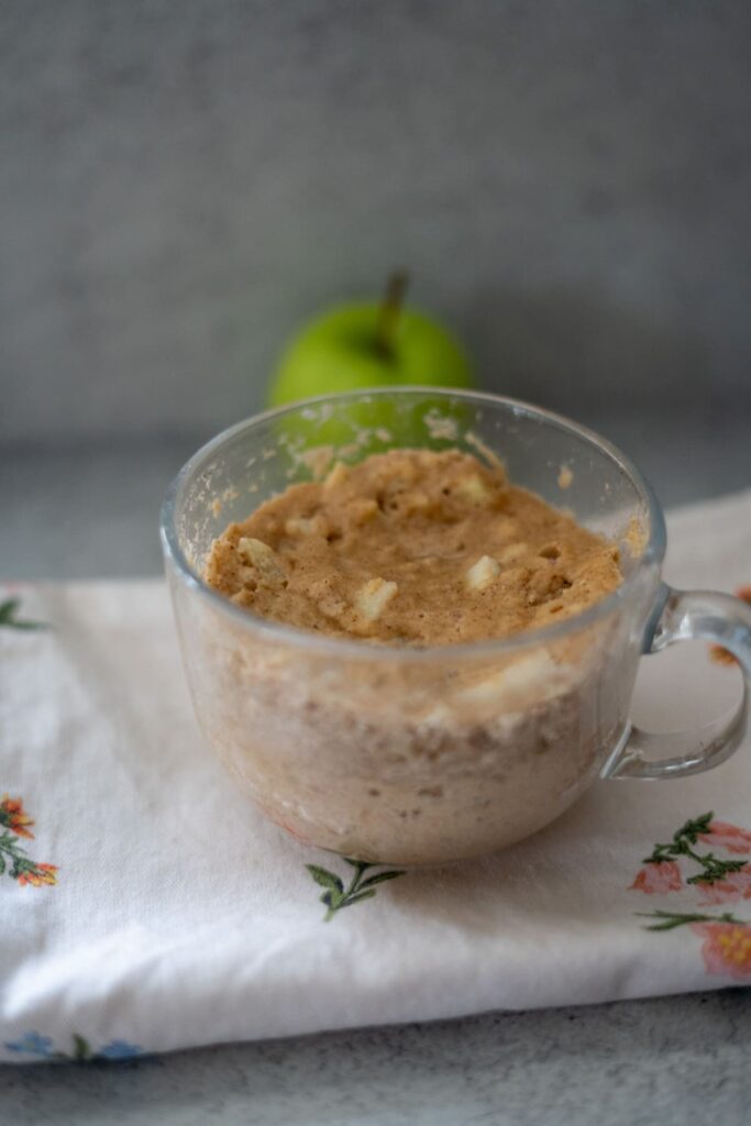 A glass mug filled with a light brown keto apple cinnamon mug cake, with small chunks visible, sits on a floral-patterned cloth. A green apple is in the background.