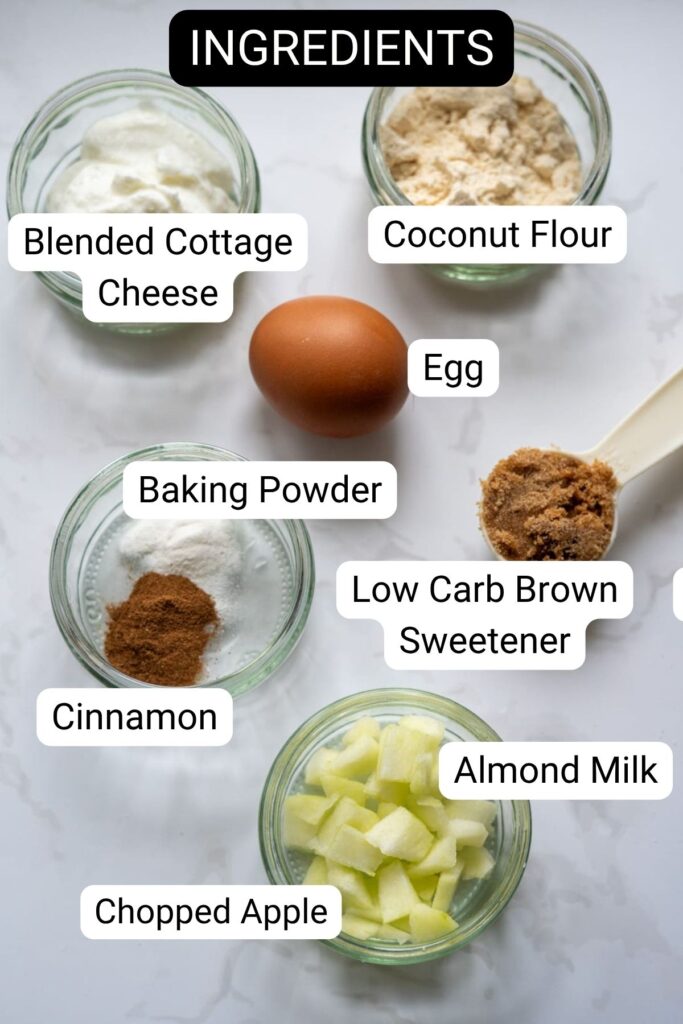 Ingredients for a keto apple cinnamon mug cake are displayed, including cottage cheese, coconut flour, an egg, baking powder, low carb brown sweetener, cinnamon, almond milk, and chopped apple in small bowls.