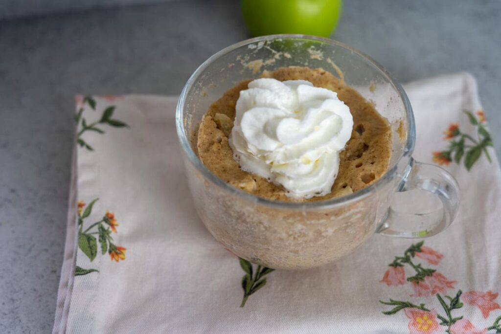 A clear glass mug filled with a light brown keto apple cinnamon mug cake topped with whipped cream, placed on a floral napkin. A green apple is partially visible in the background.