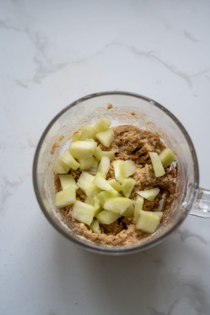 A glass mug containing a mixture of chopped green apples and batter for a keto apple cinnamon mug cake sits on a white marble surface.