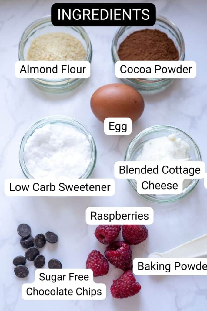 Top-down view of labeled baking ingredients on a white surface for a keto chocolate raspberry mug cake: almond flour, cocoa powder, egg, low carb sweetener, blended cottage cheese, raspberries, baking powder, and chocolate chips.