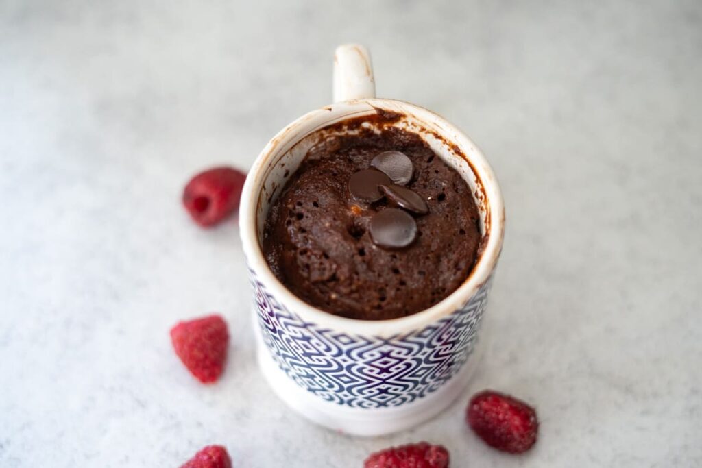 A chocolate mug cake topped with chocolate chips in a patterned mug, surrounded by a few raspberries on a light surface.