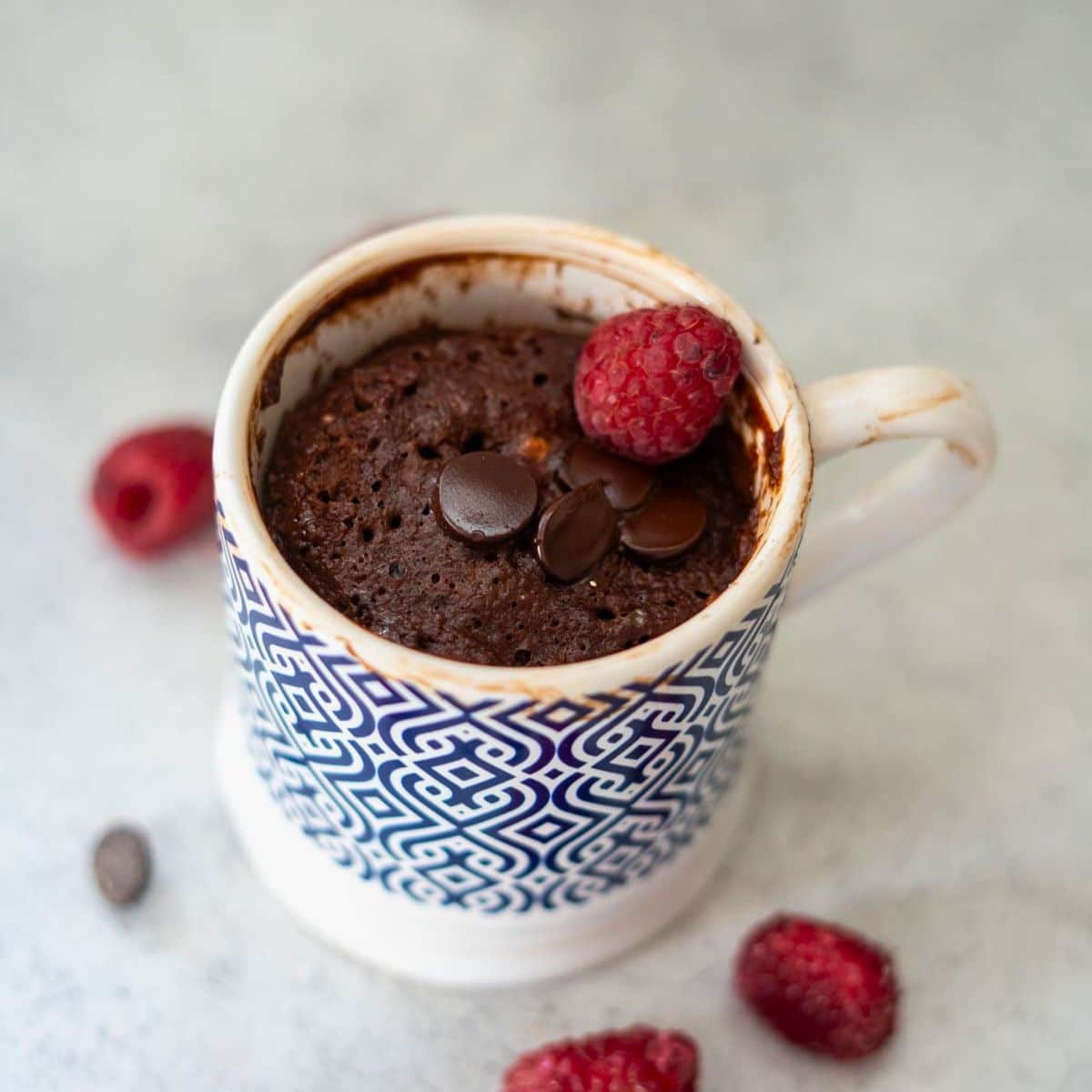 A chocolate mug cake topped with chocolate chips and a raspberry, served in a patterned mug with a few raspberries scattered nearby.