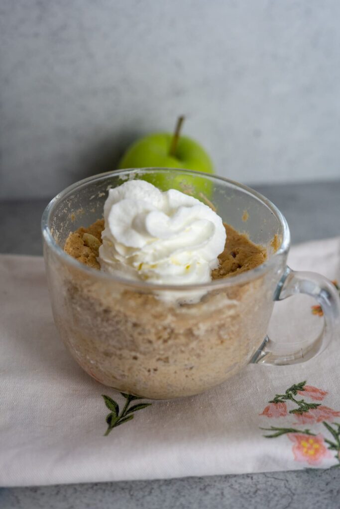 A glass mug with a keto apple cinnamon mug cake topped with whipped cream, placed on a floral napkin, with a green apple in the background.