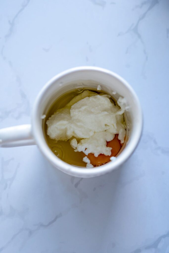 A white mug containing a raw egg and a dollop of yogurt, ready to be transformed into a keto chocolate raspberry mug cake, sits on a white marble surface.