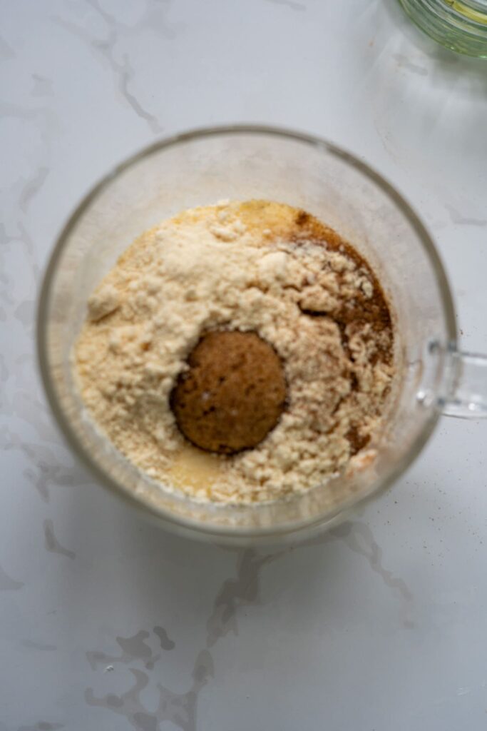 A glass mug containing flour, a brown mixture, and liquid ingredients for a keto apple cinnamon mug cake on a white marble surface, viewed from above.