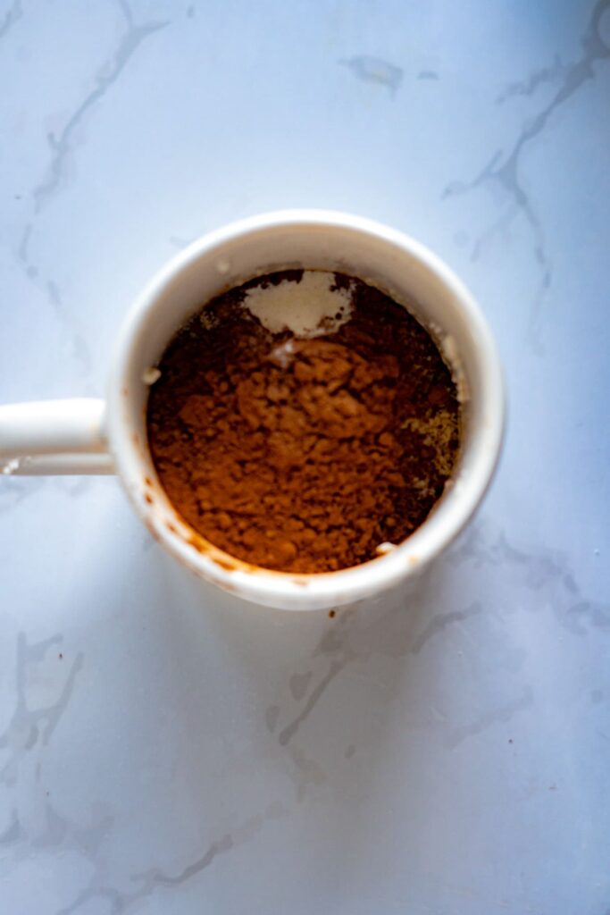 A white mug containing dry ingredients for a keto chocolate raspberry mug cake, including cocoa powder and sugar, rests on a light marble surface.