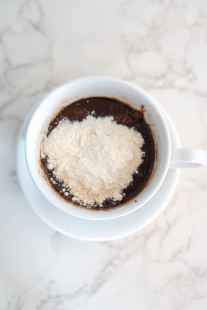 A white mug filled with chocolate batter and a pile of white flour on top, placed on a white saucer on a marble surface.