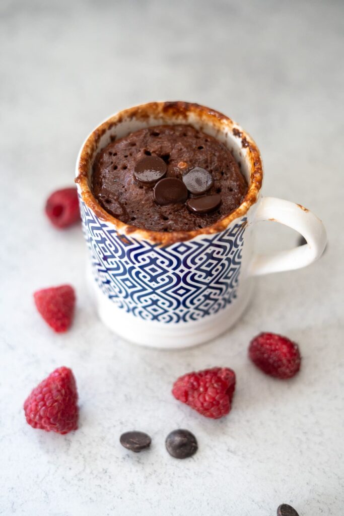A keto chocolate raspberry mug cake topped with chocolate chips in a patterned mug, surrounded by fresh raspberries and extra chocolate chips on a light surface.