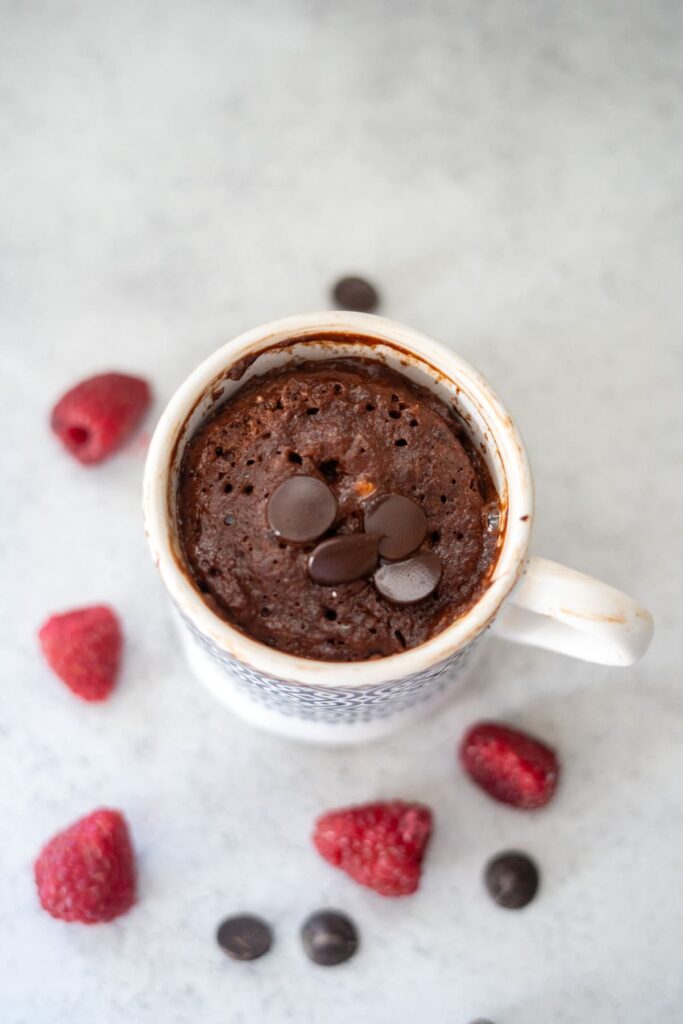 A keto chocolate raspberry mug cake topped with chocolate chips in a white mug, surrounded by scattered raspberries and chocolate chips on a light surface.