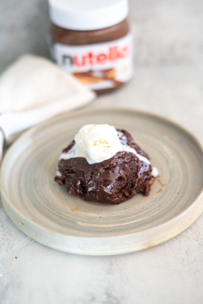 A chocolate dessert topped with a scoop of vanilla ice cream sits on a beige plate, with a jar of Nutella in the background.