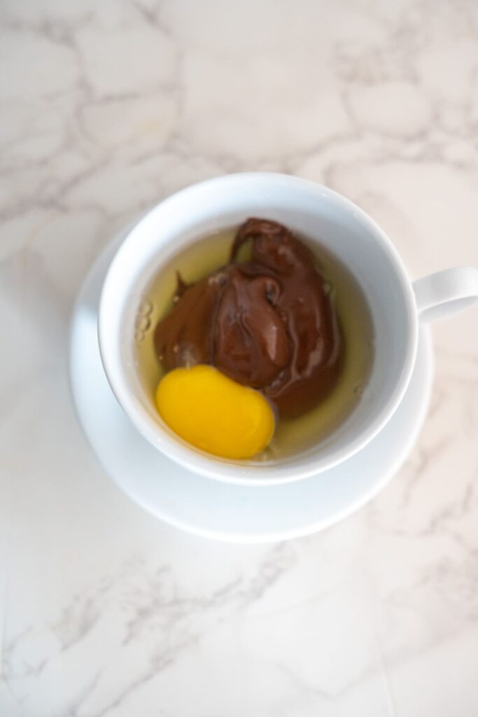 A white cup containing an egg yolk, egg white, and a dollop of chocolate spread, placed on a white saucer atop a marble surface.