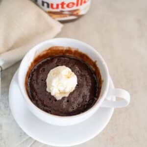 A chocolate mug cake with a scoop of vanilla ice cream on top served in a white cup and saucer, with a napkin and Nutella jar in the background.