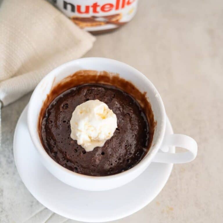A chocolate mug cake with a scoop of vanilla ice cream on top served in a white cup and saucer, with a napkin and Nutella jar in the background.
