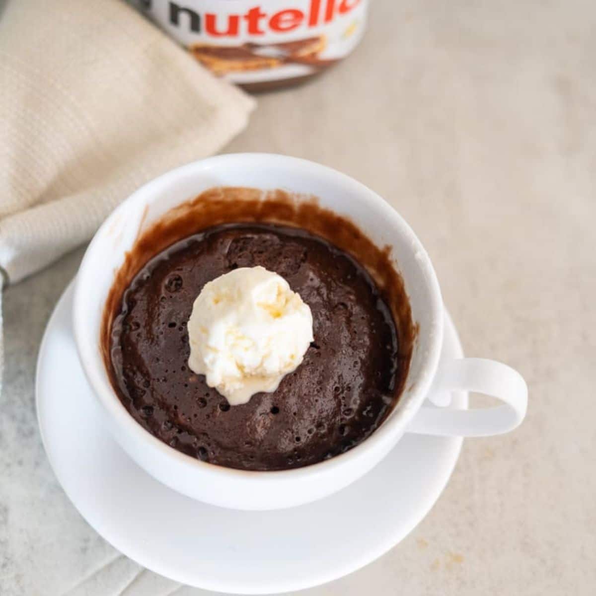 A chocolate mug cake with a scoop of vanilla ice cream on top served in a white cup and saucer, with a napkin and Nutella jar in the background.