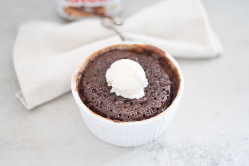 A chocolate mug cake in a white ramekin topped with a scoop of vanilla ice cream, placed in front of a folded beige napkin.