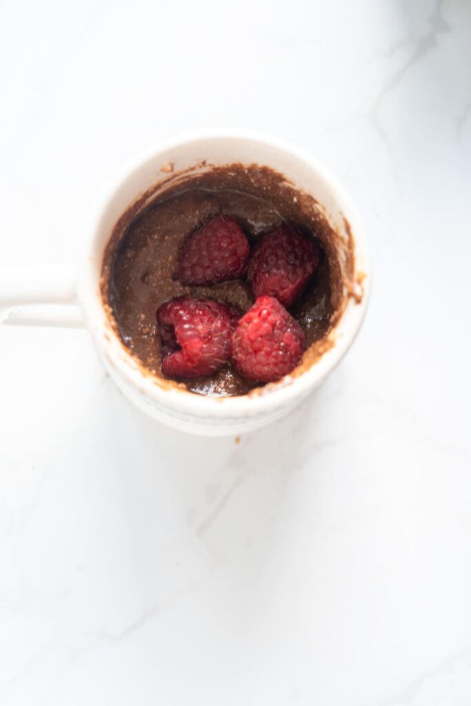 A white mug filled with keto chocolate raspberry mug cake batter and topped with fresh raspberries sits on a white surface.