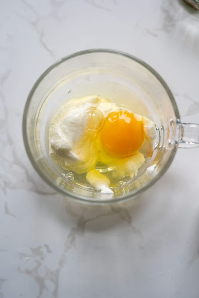 A glass mug containing a raw egg and a dollop of white yogurt or cream sits on a white marble surface, ready to be transformed into a keto apple cinnamon mug cake.