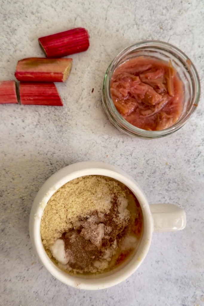 A mug filled with baking ingredients, a few pieces of raw rhubarb, and a small jar of cooked rhubarb are arranged on a light surface—perfect for making a quick keto rhubarb mug cake.