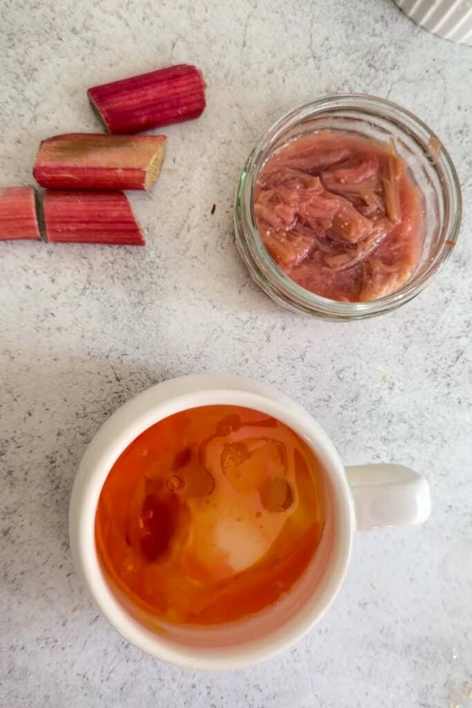 A white mug of tea with milk being poured, next to a glass jar of stewed rhubarb, several pieces of raw rhubarb, and a keto rhubarb mug cake on a light countertop.