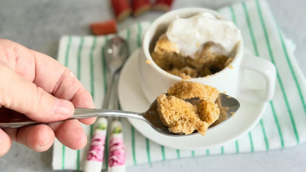 A hand holds a spoonful of mug cake topped with whipped cream, with the mug and utensils on a white plate and striped napkin in the background.