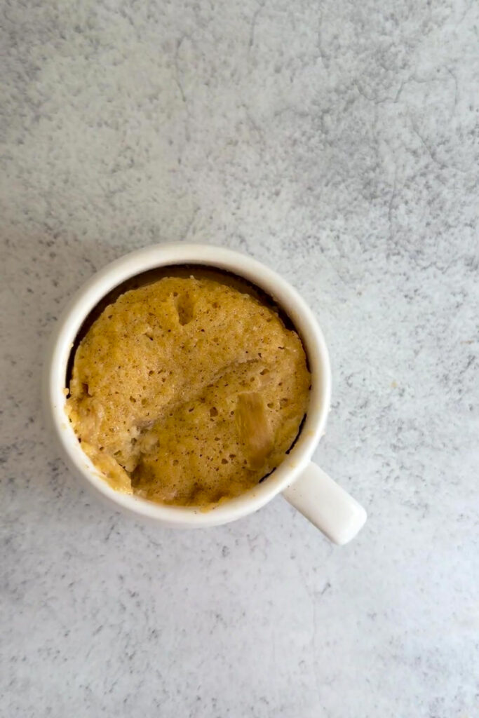 A white mug filled with a freshly baked keto rhubarb mug cake sits on a light gray textured surface.