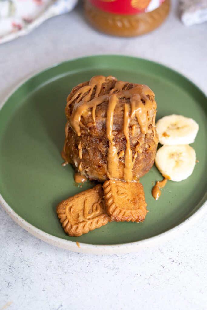 A biscoff banana mug cake topped with peanut butter drizzle, served on a green plate with banana slices and two rectangular cookies.
