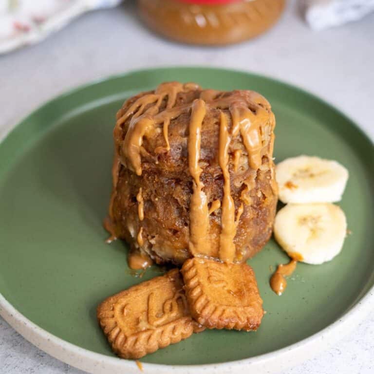A small cake drizzled with peanut butter sits on a green plate, accompanied by two round banana slices and two rectangular cookies.