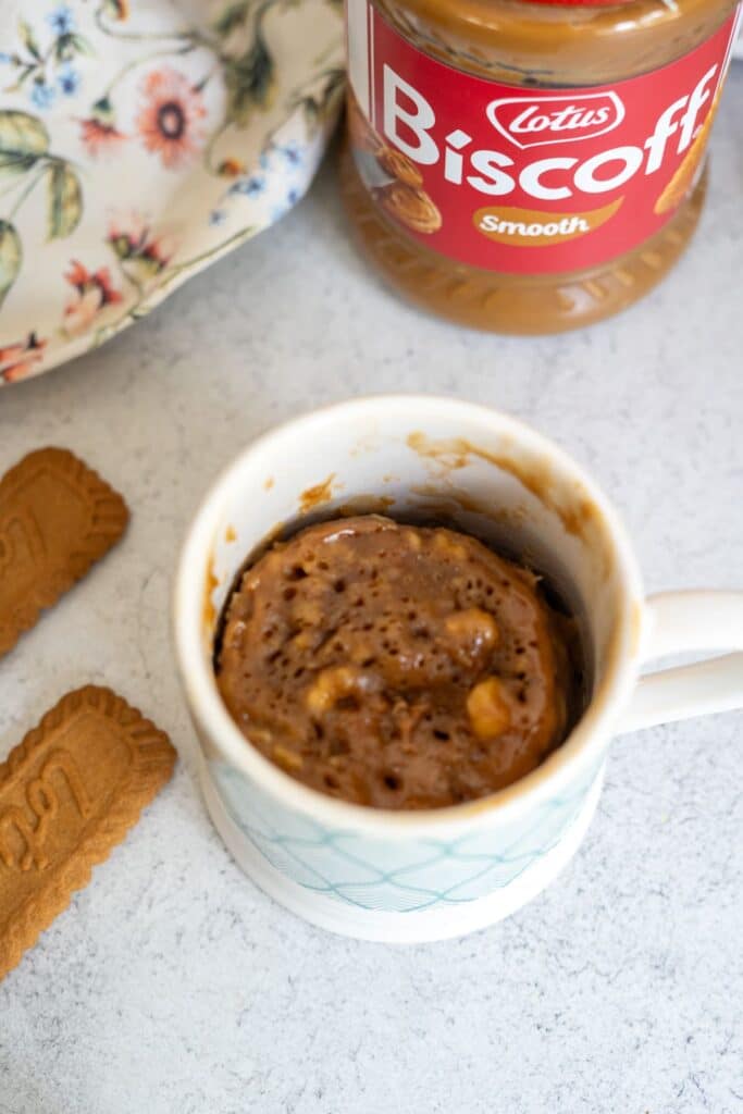 A mug cake sits in a white mug next to two Biscoff cookies and a jar of Lotus Biscoff smooth spread on a light surface.