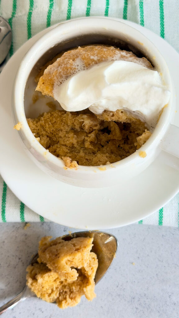 A keto rhubarb mug cake topped with whipped cream sits on a white plate, with a spoonful of cake resting on the counter beside it.