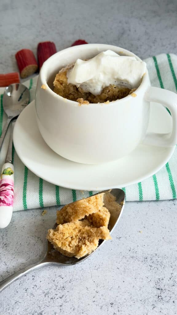 A keto rhubarb mug cake topped with whipped cream sits on a saucer, with a spoonful of cake placed beside it on a white surface.