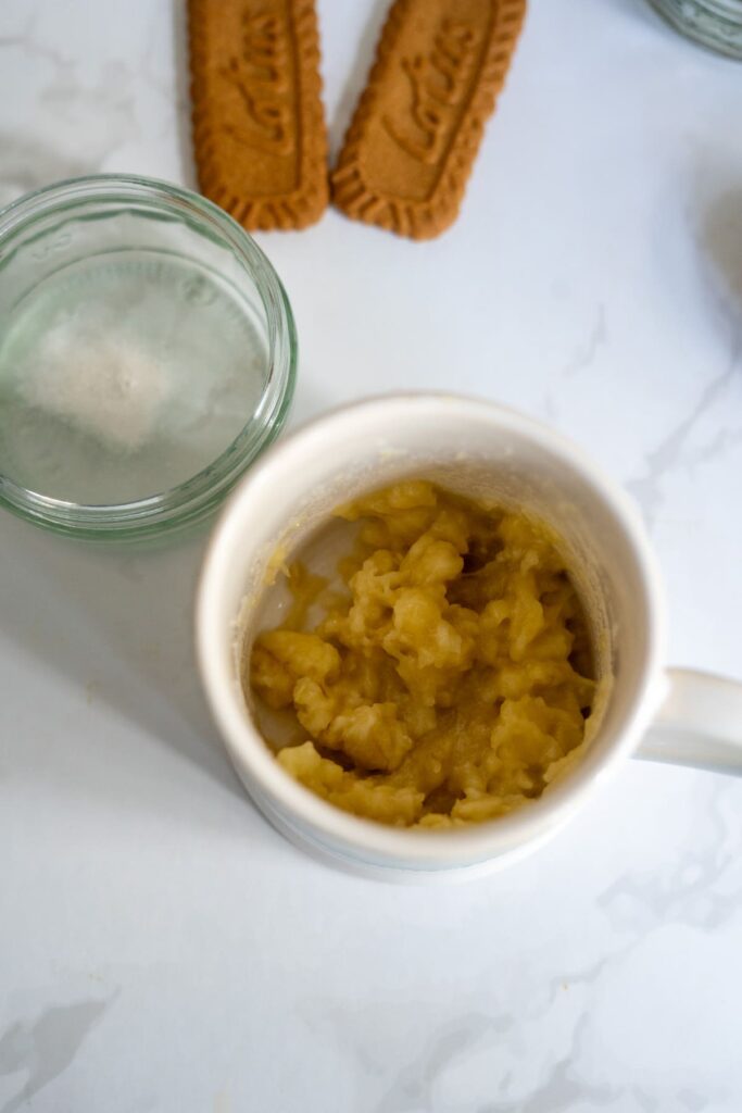 A mug containing mashed banana sits on a white surface next to a glass of water and two rectangular cookies.