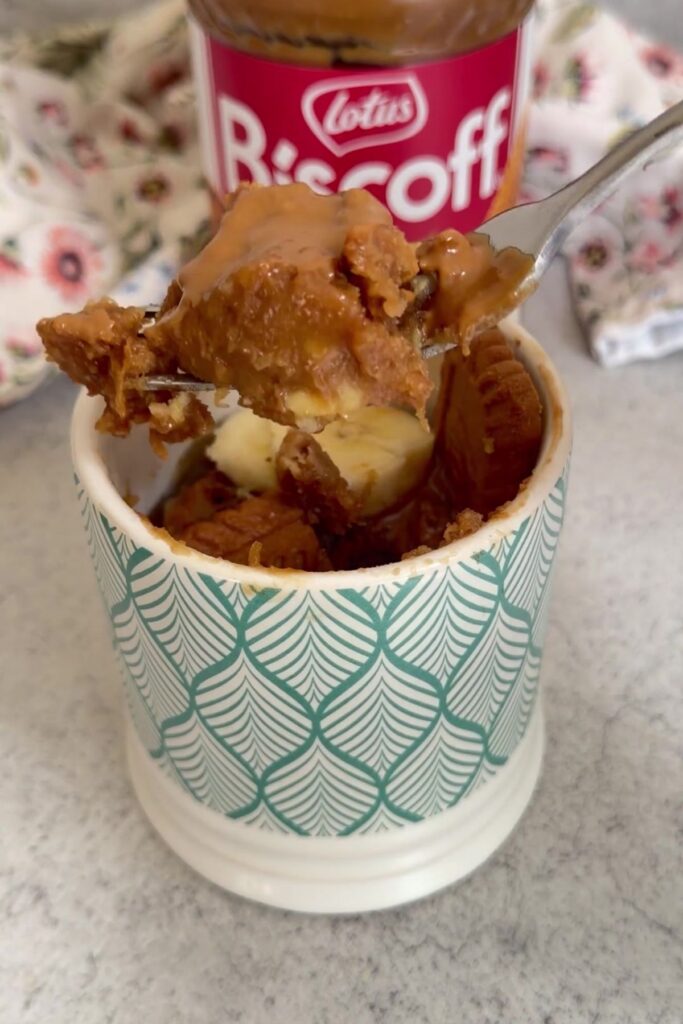 A spoonful of dessert with peanut butter and a cookie topping above a cup, with a jar of Lotus Biscoff spread in the background—perfect for enjoying a biscoff banana mug cake.
