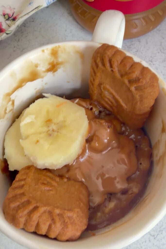 A white mug filled with oatmeal, topped with sliced banana, crumbled biscoff biscuits, and a spread of peanut butter—like a cozy biscoff banana mug cake for breakfast.