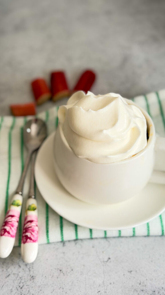 A white cup filled with a hot drink topped with whipped cream, placed on a saucer and striped towel, next to two spoons and vibrant red keto rhubarb mug cake in the background.