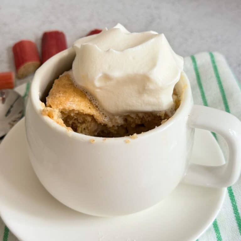A mug cake topped with whipped cream sits on a white saucer, with pieces of rhubarb in the background and a green-striped napkin beside it.