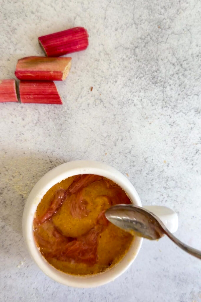 A small white bowl of custard-like keto rhubarb mug cake with rhubarb on top, being stirred with a spoon; pieces of fresh rhubarb lie beside the bowl on a light surface.