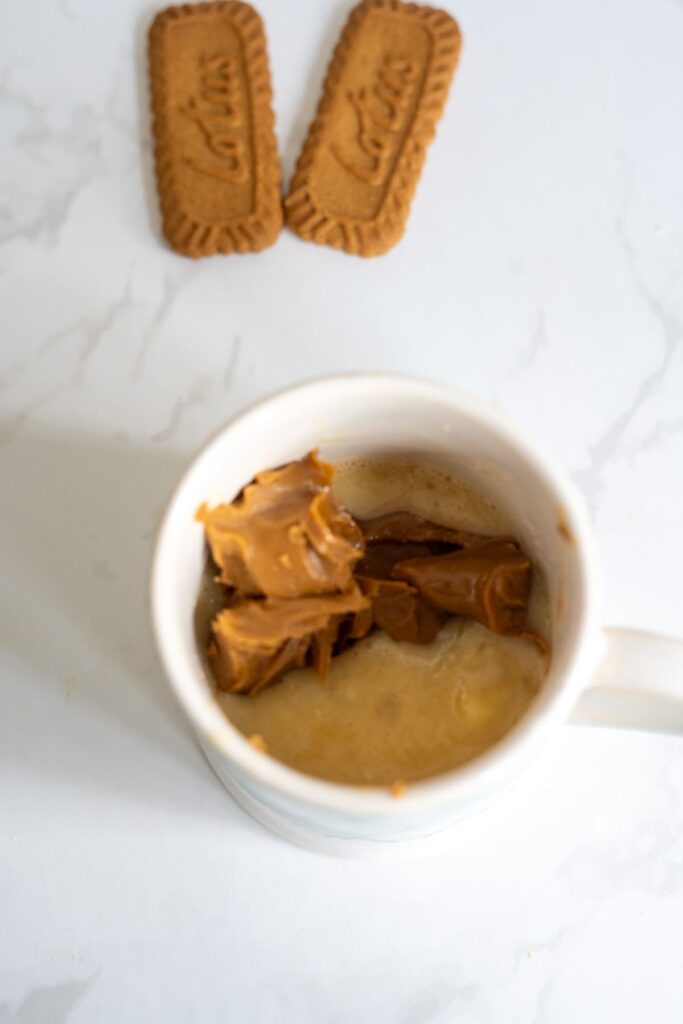A white mug containing melted butter and cookie butter, with two rectangular cookies on a white surface beside it.