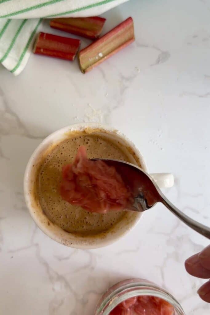 A spoonful of rhubarb compote is being added to a cup of coffee on a white countertop, with rhubarb pieces and a striped towel in the background.
