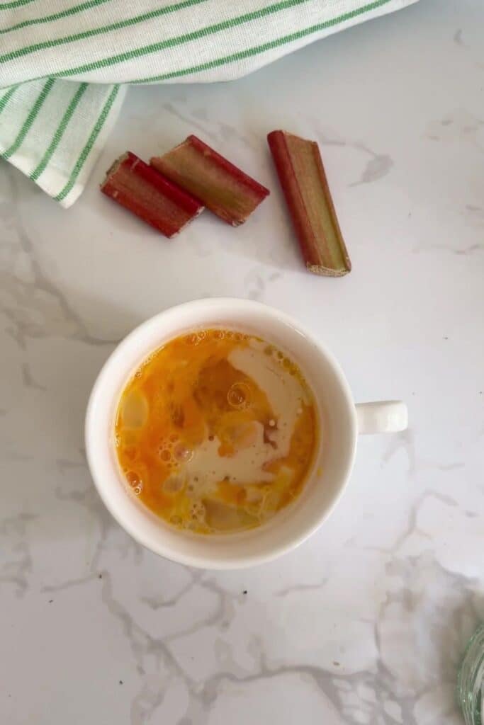 A white mug with eggs and milk inside sits on a marble countertop near sliced rhubarb and a green-striped towel.