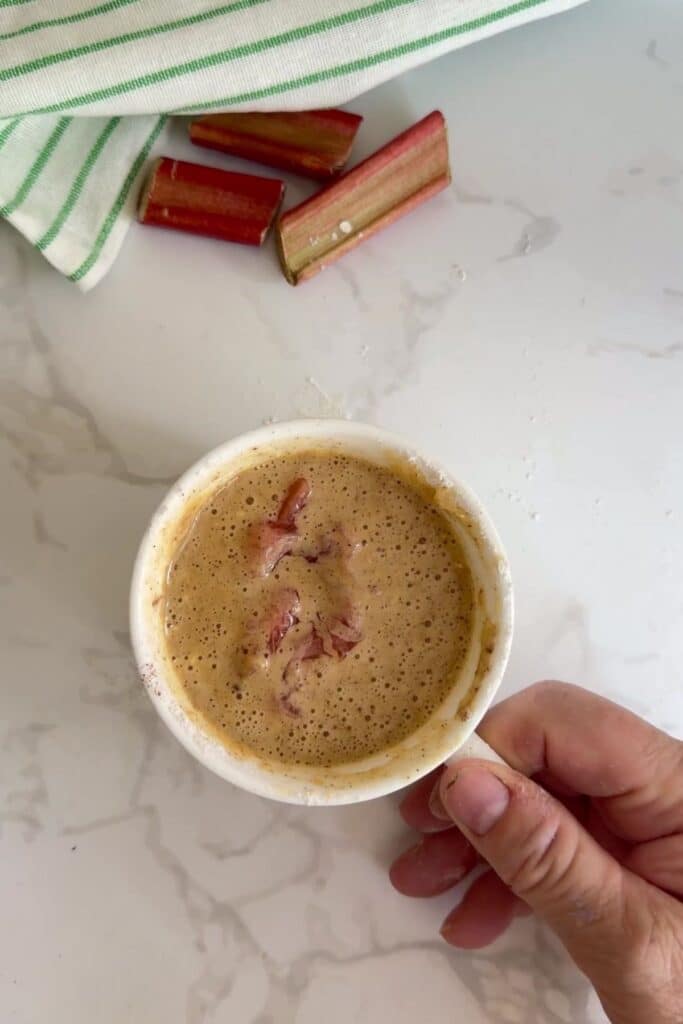 A hand holds a small white bowl filled with a frothy mixture and rhubarb pieces; cut rhubarb and a striped cloth are in the background on a marble surface.