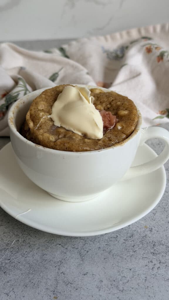 A mug cake topped with a dollop of cream sits in a white cup on a matching saucer, with a floral cloth in the background.