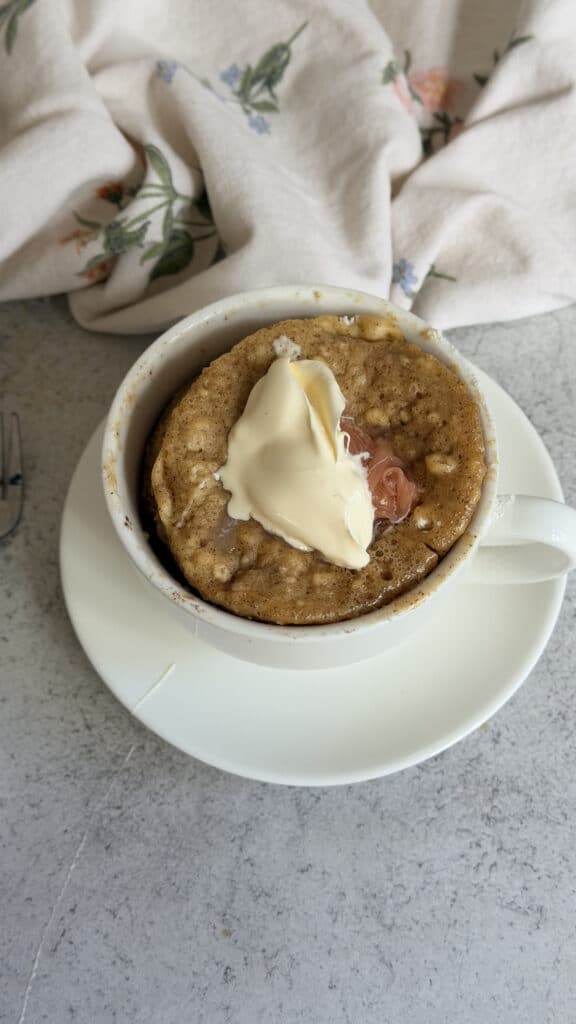 A mug cake topped with a dollop of cream and fruit preserves sits on a white saucer, with a floral cloth in the background.