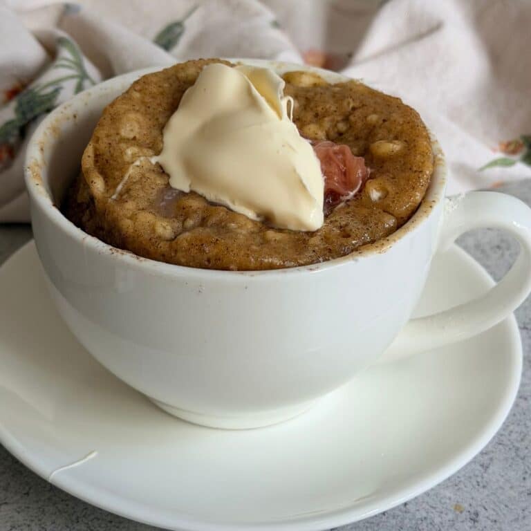 A white cup on a saucer holds a baked mug cake topped with a dollop of cream and a visible spot of fruit filling, set against a neutral background.