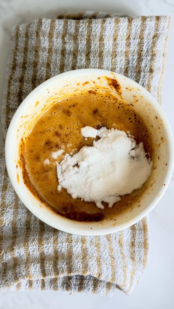 A white bowl containing an almond butter mug cake mixture topped with a mound of white powder, resting on a striped kitchen towel.