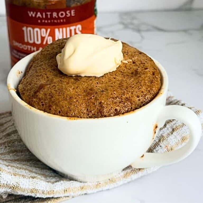 A mug cake topped with a dollop of cream, with a jar of Waitrose peanut butter in the background.