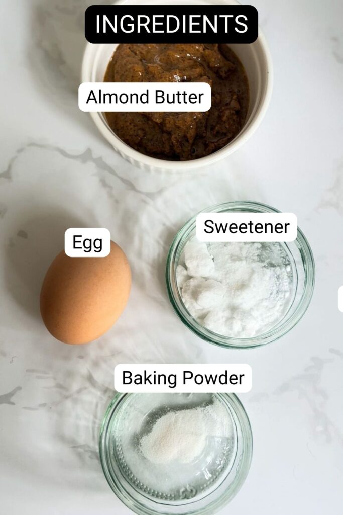 Ingredients for an almond butter mug cake: a bowl of almond butter, one egg, a small dish of sweetener, and a small dish of baking powder.